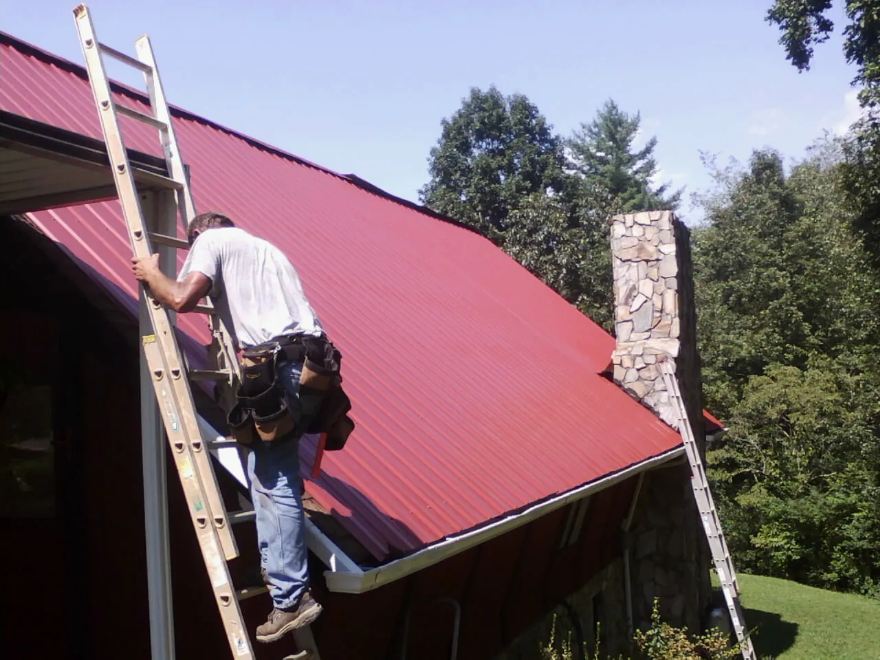 Roofer on a ladder beside a red metal roof and stone chimney — Soffit & Fascia Repair by Roofing Brothers in Marshall